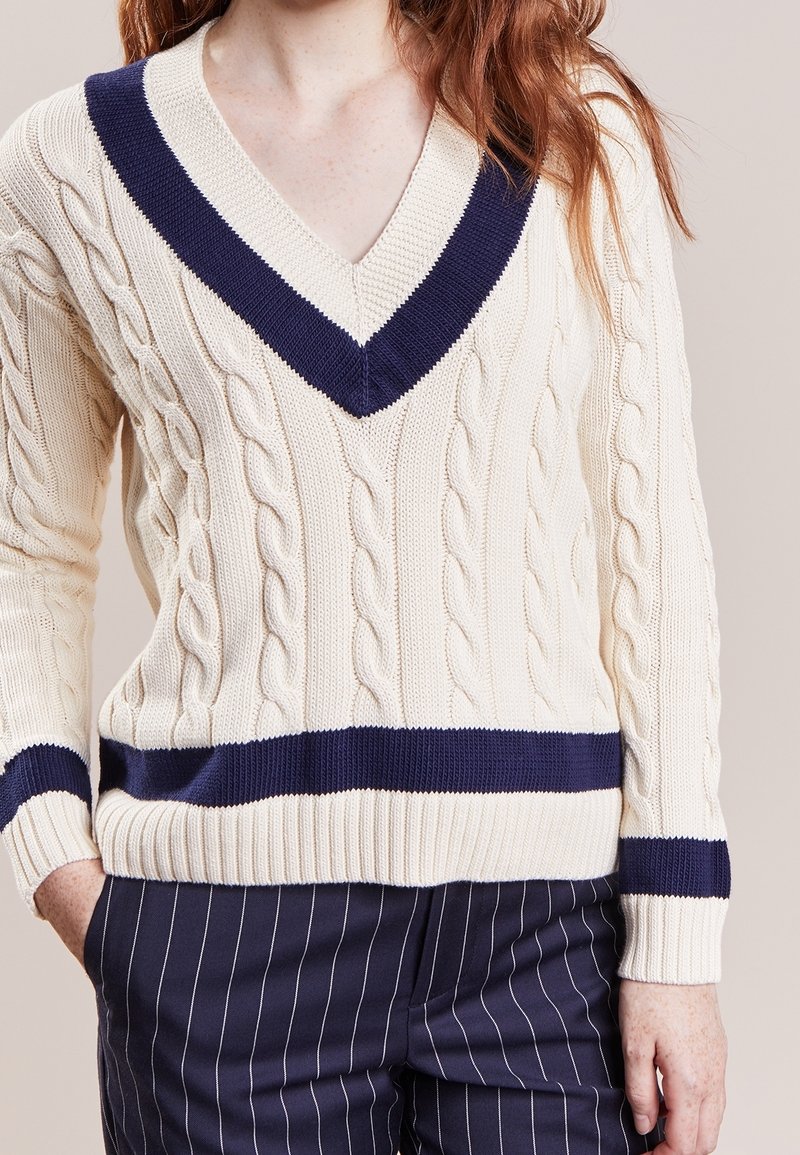 Femme portant un pull en maille torsadée crème avec un col en V bleu marine et des rayures, associé à un pantalon rayé bleu marine sur un fond beige.