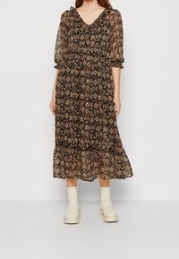 Robe midi imprimée florale noire avec manches bouffantes, ourlet à volants et taille élastique, présentant un mélange de motifs complexes aux tons naturels. Portée avec des bottes beiges.