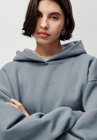 Une jeune femme aux cheveux courts et foncés porte des boucles d'oreilles en argent et un sweat à capuche bleu clair, les bras croisés, contre un fond uni.