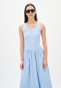 Robe bleu clair avec un corsage à côtes et une jupe froncée, assortie de lunettes de soleil cat-eye noires. Texture lisse, design sans manches.