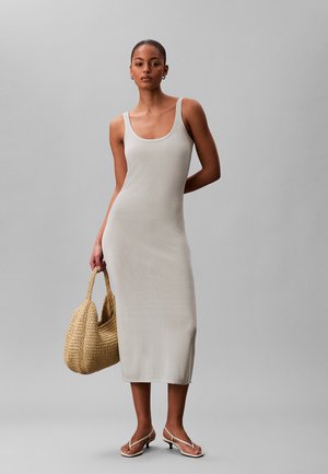 Mujer vistiendo un vestido midi sin mangas de color gris claro con sandalias blancas de tacón, sosteniendo un bolso grande de mimbre tejido contra un fondo gris liso.