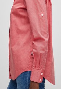 Langærmet koralrosa skjorte med knapmanchetter, båret over blå jeans, som viser side søm og stof tekstur detaljer.