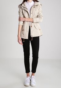Veste légère beige avec capuche, taille à cordon de serrage et multiples poches, associée à un pantalon noir ajusté et des baskets blanches.