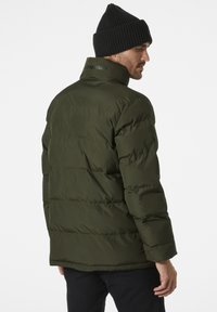 Groene pufferjack met horizontale stiksel, hoge kraag en zichtbaar logo op de achterkant. Draagt een zwarte gebreide beanie.