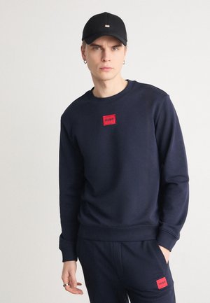 Marinblå sweatshirt med en röd "HUGO" logotyp patch, ribbad mudd och nederkant, parat med matchande byxor, under en enkel svart keps.