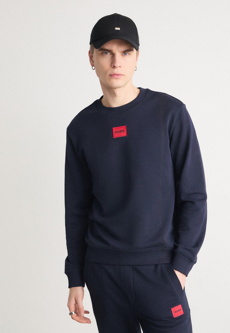 Felpa blu navy con un'etichetta rossa "HUGO", polsini e orlo a coste, abbinata a pantaloni coordinati, indossata sotto un cappellino nero semplice.