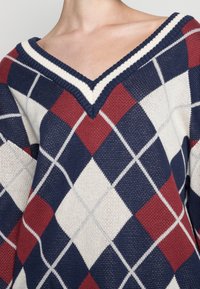 Pull en maille à col en V présentant un motif argyle géométrique en bleu marine, rouge et blanc. Texture douce avec un col côtelé contrastant.