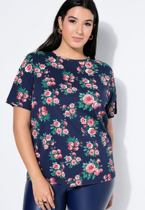 Frau mit langen dunklen Haaren, die goldene Creolen trägt und ein marineblaues Kurzarmhemd mit pinkem und grünem Blumenmuster trägt, steht vor weißem Hintergrund.