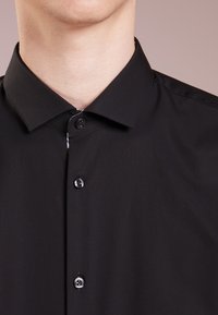 Mann trägt ein schwarzes Hemd mit Knöpfen und klassischem Kragen, gezeigt vom Hals bis zur oberen Brust vor einem einfarbigen Hintergrund.