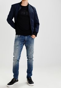 Námořnicky modrá bombardérská bunda přes černé tričko s kulatým výstřihem, roztrhané světle modré džíny a černé tenisky s bílými podrážkami, s hladkou texturou.
