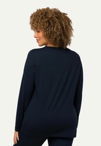 Frau mit lockigem Haar, die ein langärmliges, dunkelblaues Oberteil und passende Hosen trägt und seitlich vor einem hellen, schlichten Hintergrund steht.