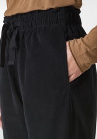 Černé kordové kalhoty s elastickým pasem a šňůrkou, obsahující boční kapsy a měkkou texturu. Vrchní část je hnědá a má dlouhé rukávy.