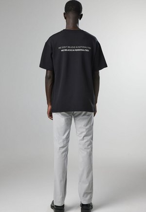 Mann, der mit dem Rücken zur Kamera steht und ein schwarzes T-Shirt mit dem Text "We don't believe in nationalities we believe in personalities" in weißer Schrift trägt.