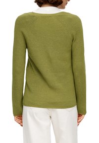Groene gebreide trui, lange mouwen, geribbelde textuur, ronde hals en schouderdetails. Draagt over lichte broek.