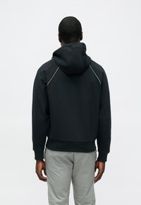 Μαύρο φούτερ με κουκούλα και κορδόνι, υφασμάτινη υφή, μανίκια ράγκλαν και αντίθετες ραφές σε λευκό χρώμα. Συνδυασμένο με ανοιχτό γκρι παντελόνι.