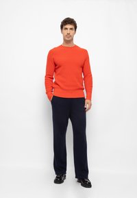 Oranssi neulepaita, jossa on uurteinen pinta, yhdistettynä tummansinisiin leveälahkeisiin housuihin ja mustiin kenkiin. Malli seisoo valkoista taustaa vasten.