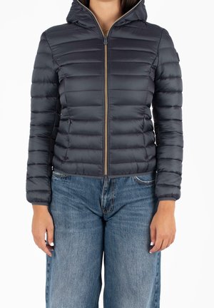 Frau trägt eine taillierte, marineblaue gesteppte Jacke mit Kapuze und goldenem Reißverschluss, kombiniert mit hoch taillierten, weit geschnittenen blauen Jeans.