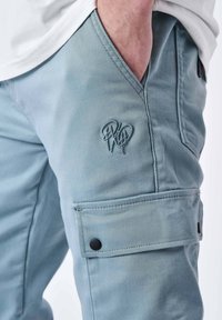 Lichtblauwe cargobroek van zachte stof met zijzakken, een knopenflap en een geborduurd logo op de dij.