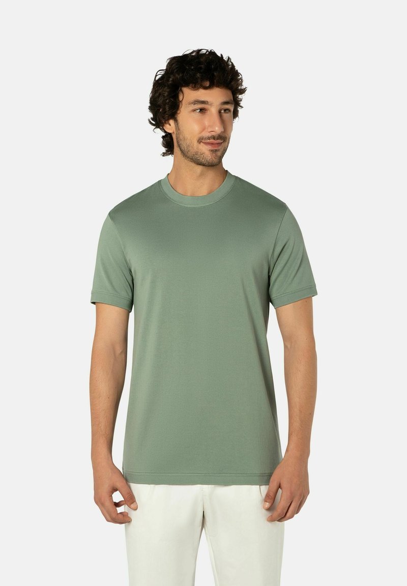 Mann mit lockigem Haar, der ein kurzärmeliges, salbeigrünes T-Shirt mit Rundhalsausschnitt und weiße Hose trägt, vor einem schlichten hellen Hintergrund steht.