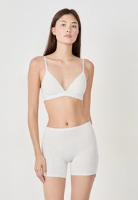 Ung kvinna som står mot en enfärgad bakgrund, klädd i en ljusgrå mjuk BH och matchande höga shorts, med armarna avslappnade längs sidorna.