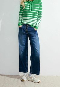 Sweat à capuche à rayures vertes et blanches, jean large bleu à braguette boutonnée, et baskets claires, présentés sur un fond neutre.