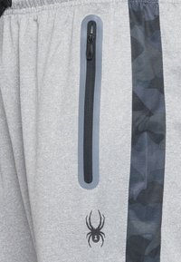 Los pantalones cortos atléticos grises presentan un bolsillo con cremallera de color negro. Un panel de camuflaje oscuro adorna el costado y se puede ver un logo de araña negro.