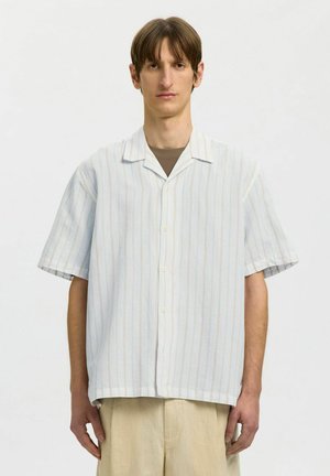 Mann iført en løs, kortermet hvit skjorte med tynne vertikale striper over en brun undertrøye og beige bukser mot en ensfarget bakgrunn.