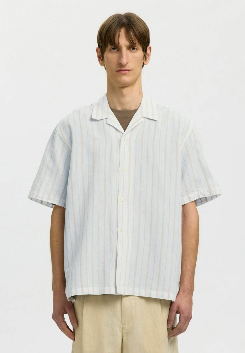 Muž v ľahkej bielej košeli s krátkymi rukávmi s tenkými vertikálnymi pruhmi, pod ňou hnedé tričko a béžové nohavice, na jednoduchom pozadí.