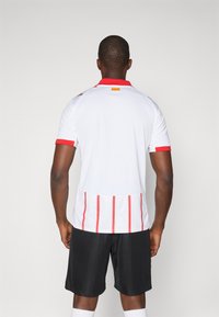 Camisa desportiva branca com detalhes vermelhos, mangas curtas e riscas verticais vermelhas nas costas. Combinada com calções pretos e meias brancas.