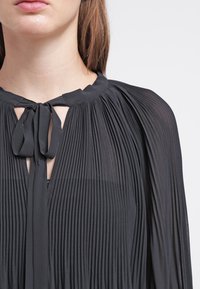 Czarna, plisowana bluzka z wiązaniem przy szyi, wykonana z lekkiego, zwiewnego materiału, z głębokim dekoltem w kształcie litery V. Teksturowany i drapowany design.
