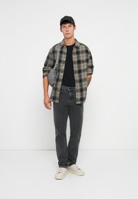 Camisa de franela a cuadros en gris y verde, camiseta negra, vaqueros grises, zapatillas blancas y una gorra negra. Bolso bandolera en gris con una correa.