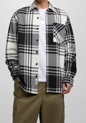 Camisa de cuadros en blanco y negro con diseño de botones, que cuenta con un bolsillo en el pecho y patrones de manga en contraste. Lleva encima una camiseta blanca.