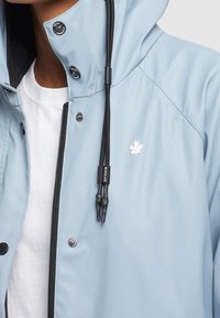 Giacca impermeabile azzurro chiaro con zip e chiusura a scatto, caratterizzata da un logo bianco a forma di foglia, accessori neri e cordini regolabili.
