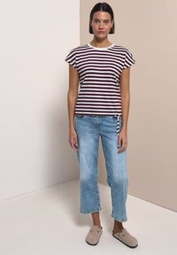 Femme debout portant un haut à manches courtes rayé blanc et bordeaux, un jean court bleu clair et des chaussures beige sans lacets.