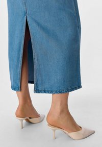 Jupe en denim avec une fente latérale, de couleur bleue, longueur mi-mollet. Associée à des mules beiges à bout pointu sur un talon fin, montrant un design minimaliste.