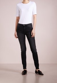 Jeans negros de corte ajustado, elaborados en denim, con una cintura de altura media, cinco bolsillos y cierre frontal de botón. Combinados con una blusa blanca de manga corta.