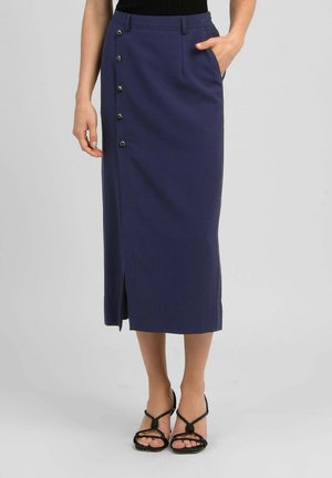 Femme portant une jupe midi bleu marine avec cinq boutons sur un côté et une fente devant, associée à des sandales à talons noires à brides.
