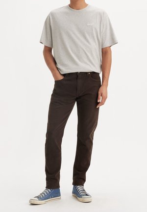 Persona con camiseta gris claro de Levi's, jeans marrón oscuro y zapatillas altas azules, de pie con una mano en el bolsillo.