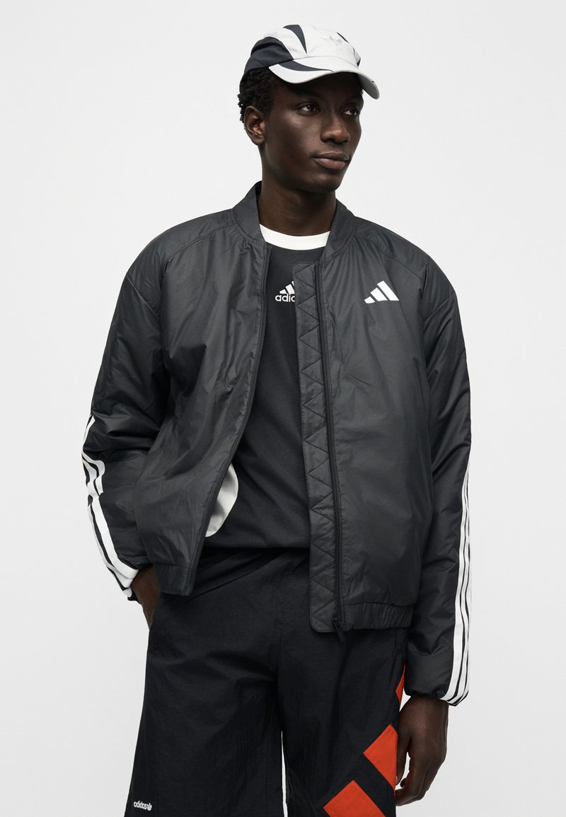 Schwarze, leichte Bomberjacke mit weißen Streifen an den Ärmeln, vorne mit Reißverschluss und gesteppter Optik. Wird über einem schwarzen Shirt und schwarzen Shorts getragen.