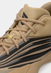 Μπεζ αθλητικό παπούτσι με υφή στο πάνω μέρος, μαύρες ρίγες ως διακοσμητικά στοιχεία και κορδόνια σε αντίθετο χρώμα. Σημαντικές λεπτομέρειες περιλαμβάνουν ένα λογότυπο και μια κίτρινη ένδειξη.