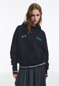 Fekete kapucnis pulóver elülső zsebbel, fehér és türkiz színű felirattal. Szürke réteges szoknya fölött, vízszintes csíkos részletekkel viselve.