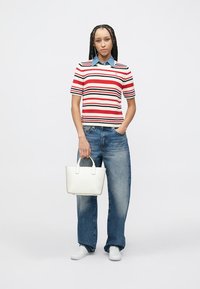 Ριγέ πλεκτό πόλο μπλουζάκι σε κόκκινο, άσπρο και ναυτικό μπλε· συνδυασμένο με φαρδιά μπλε τζιν. Κρατάει μια μικρή λευκή τσάντα χειρός· στα πόδια λευκά αθλητικά παπούτσια.