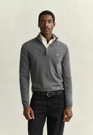 Mann trägt einen grauen Viertel-Reißverschluss-Pullover über einem weißen Hemd mit Kragen, schwarze Hose und schwarzen Gürtel vor einem schlichten hellen Hintergrund.