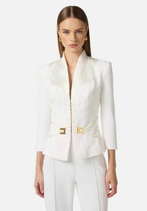 Donna che indossa un blazer bianco su misura con revers in raso, dettagli con fibbia dorata, pantaloni bianchi abbinati e orecchini quadrati dorati.