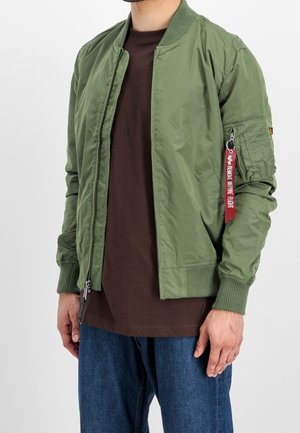 Mand iført en olivengrøn bomberjakke med en rød "Remove Before Flight"-mærkat på venstre armryglomme, en brun skjorte og blå jeans.
