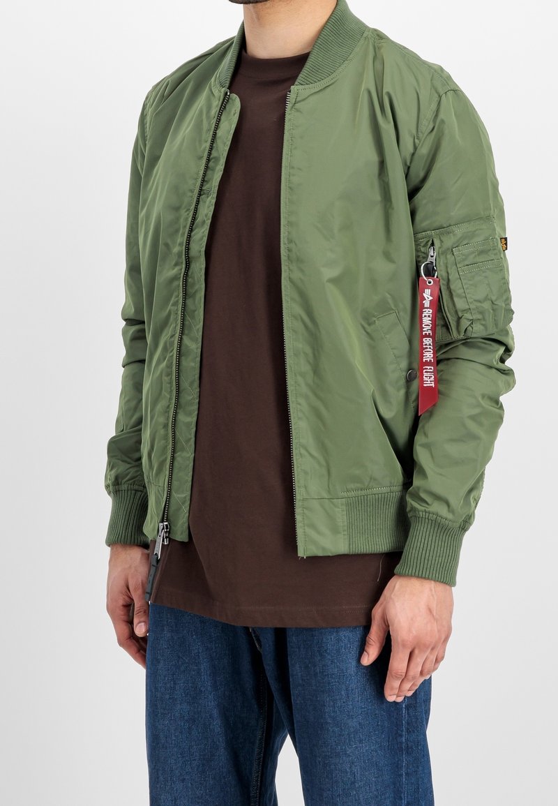 Homme portant une veste bomber vert olive avec une étiquette rouge "Remove Before Flight" sur la poche du bras gauche, une chemise marron et un jean bleu.