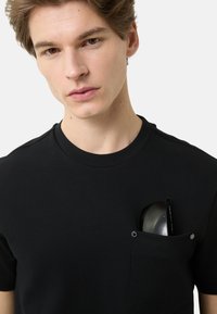 Czarna koszulka z krótkim rękawem o okrągłym dekolcie, wykonana z miękkiego materiału, z kieszenią na piersi zapinaną na srebrny zatrzask oraz częściowo widocznymi okularami przeciwsłonecznymi.