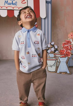 Junger Junge lacht mit den Händen in den Taschen, trägt ein hellblaues Polohemd mit Hund- und Busmotiven, braune Hose, steht neben marktthematischer Dekoration.