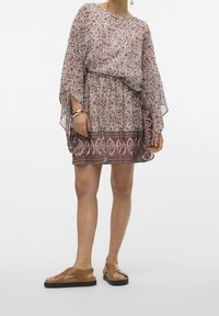 Kukkakuvioitu, puoliläpinäkyvä mekko leveillä hihoilla, joustavalla vyötäröllä ja kontrastivärisellä helmareunuksella. Päällä ruskeat tasapohjaiset sandaalit.