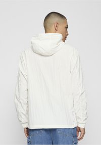 Balta kapuce ar teksturētu apdari, vaļīgs piegriezums un elastīgas aproces, kopā ar gaiši zilām džinsa šortām.